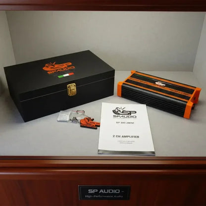 SP Audio SP 300 Mini 2-chamber amplifier shown with its black branded storage box, orange and black amplifier casing, small hardware kit, and printed instruction booklet on a shelf.