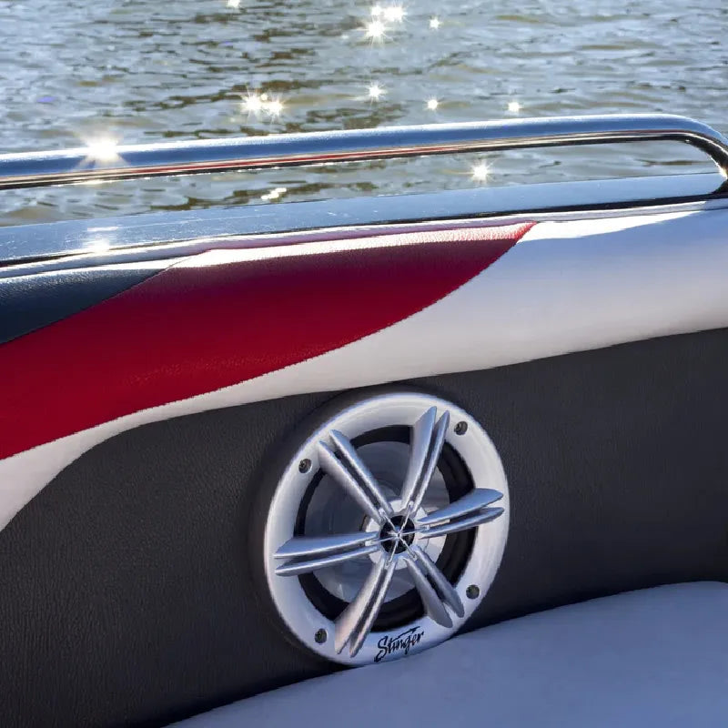 A close-up of a boat's side seating area featuring a round chrome and silver speaker with a black surround, mounted on a black and red upholstered panel. The background shows water with bright sunlight reflections.