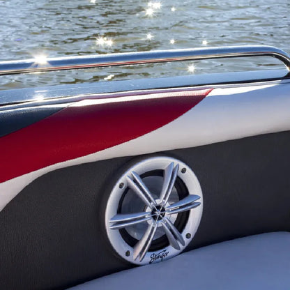 A close-up of a boat's side seating area featuring a round chrome and silver speaker with a black surround, mounted on a black and red upholstered panel. The background shows water with bright sunlight reflections.