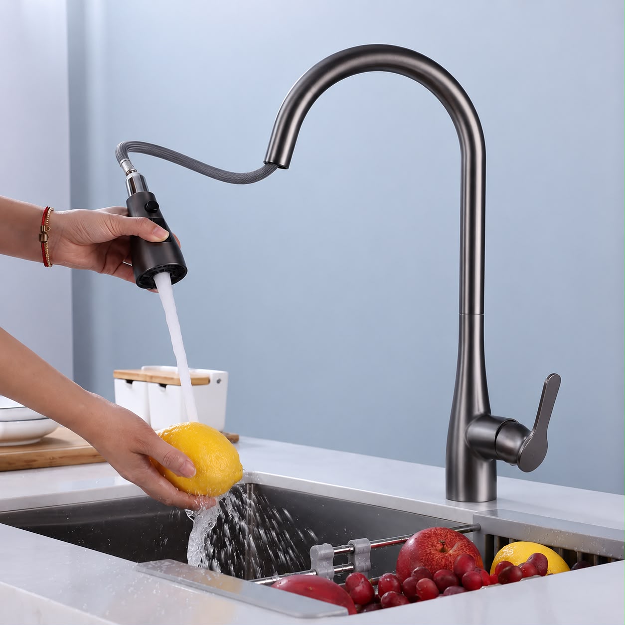 A modern matte black gooseneck faucet with a pull-out sprayer being used to rinse a yellow citrus fruit over a stainless steel sink, with fruit and a white container in the background.